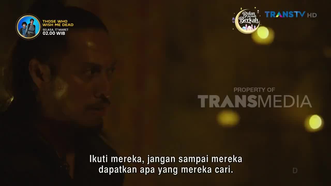 A man with a ponytail looks intently to his left, his face partially obscured by shadow. Behind him, yellow lights glow softly, hinting at a night scene in Indonesia.