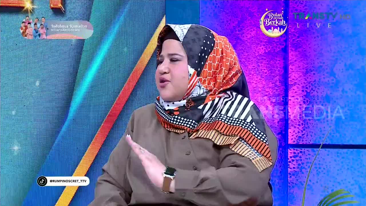 A woman in a colorful hijab speaks animatedly, her hand gesturing as she talks on a Trans TV broadcast. The backdrop behind her features bright, abstract patterns and the Trans TV logo. A woman in a colorful hijab speaks animatedly, her hand gesturing as she talks on a Trans TV broadcast. The backdrop behind her features bright, abstract patterns and the Trans TV logo.