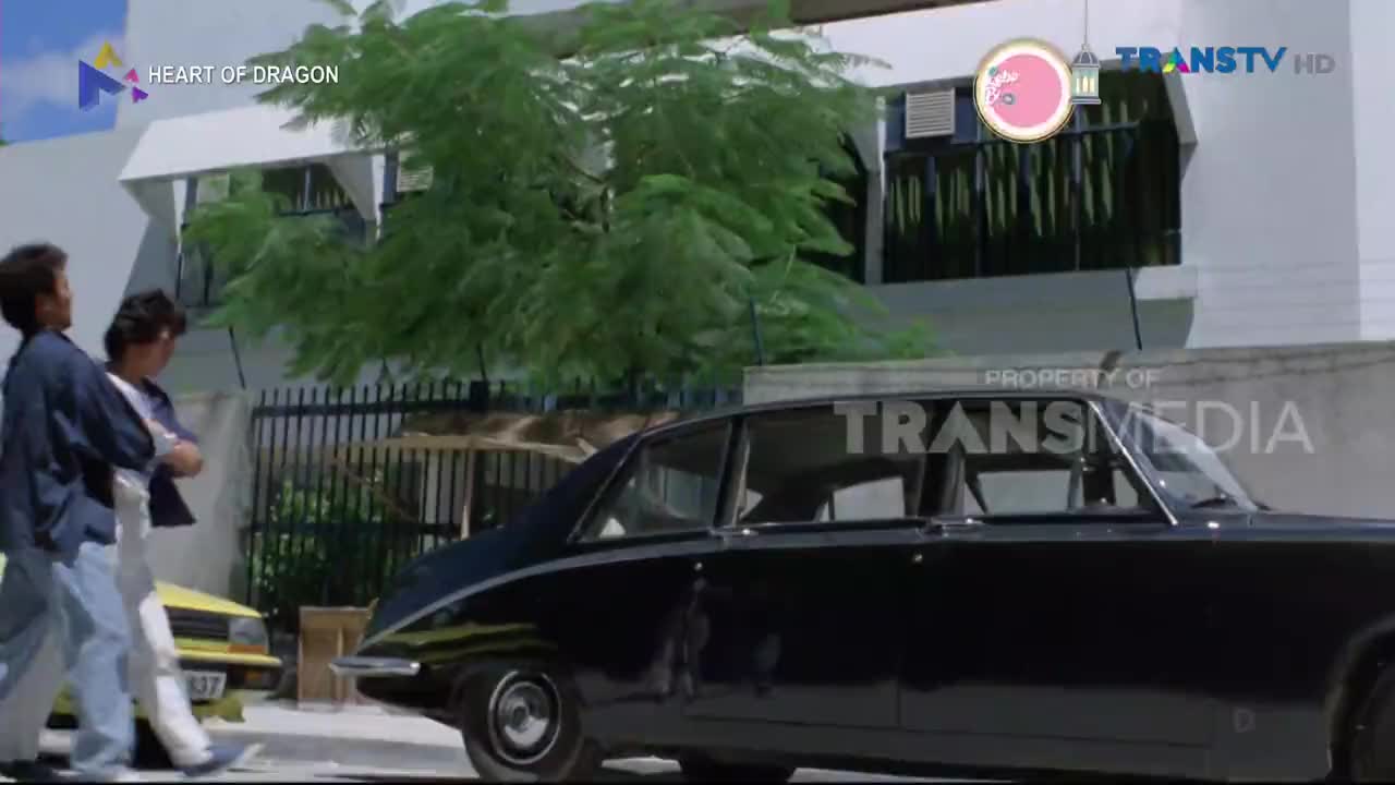 Two men in casual shirts and jeans walk past a parked black sedan. A yellow car is visible behind them, parked near a building with lush green trees.