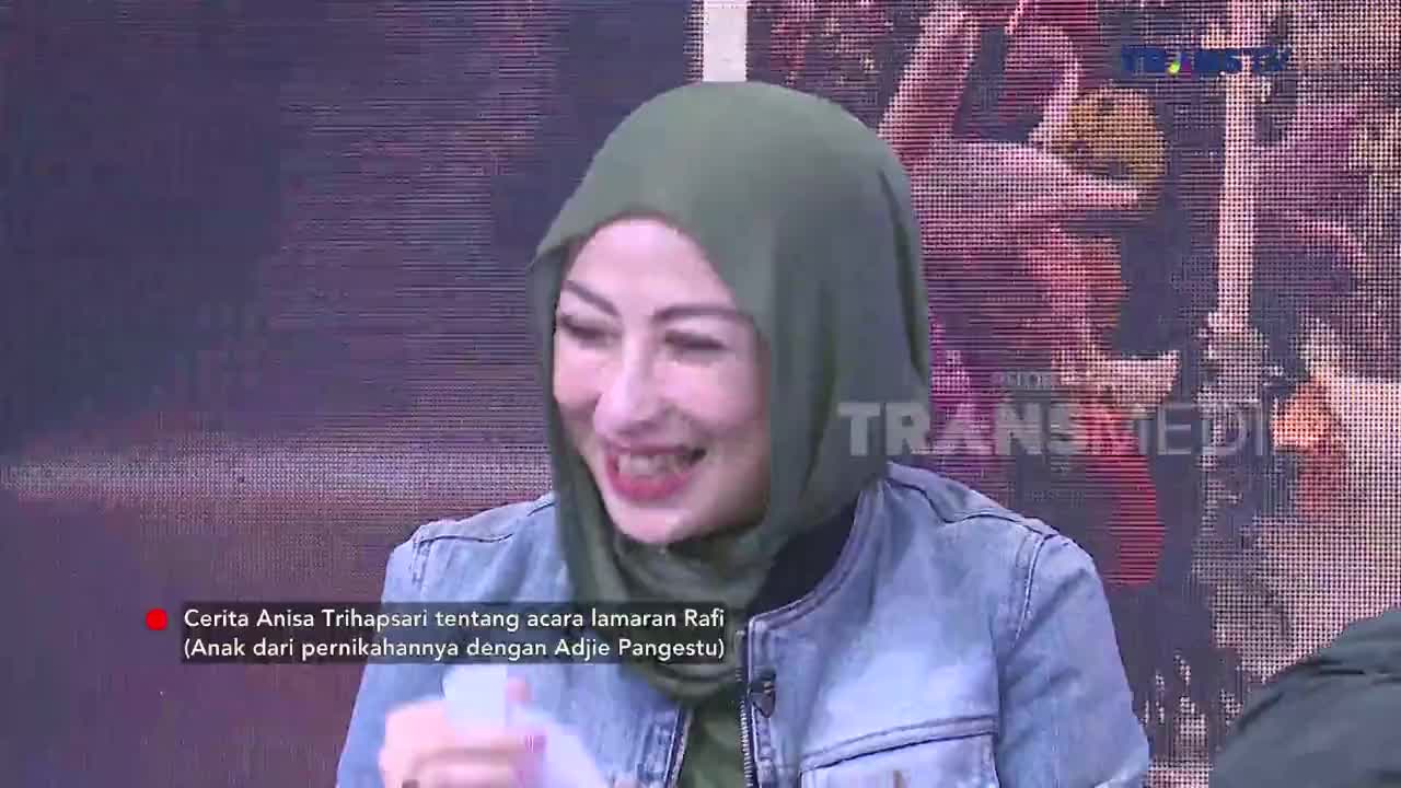 A woman in a green hijab laughs heartily, her mouth open wide. She holds a white object in her left hand, her fingers gripping it tightly.