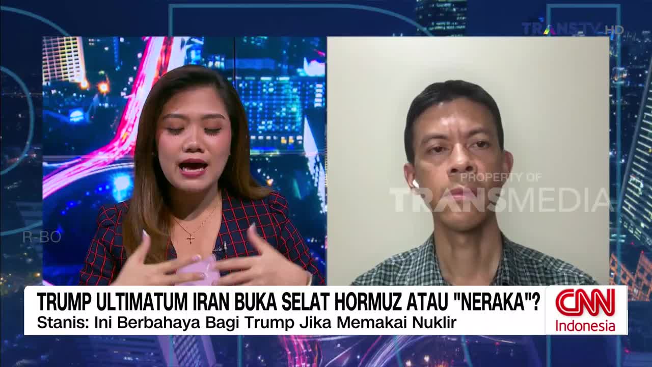 A woman with long brown hair, wearing a red and blue plaid jacket, speaks animatedly with her hands gesturing. To her right, a man with short dark hair and a checkered shirt listens, wearing white earbuds. The Trans TV logo is visible on the screen.