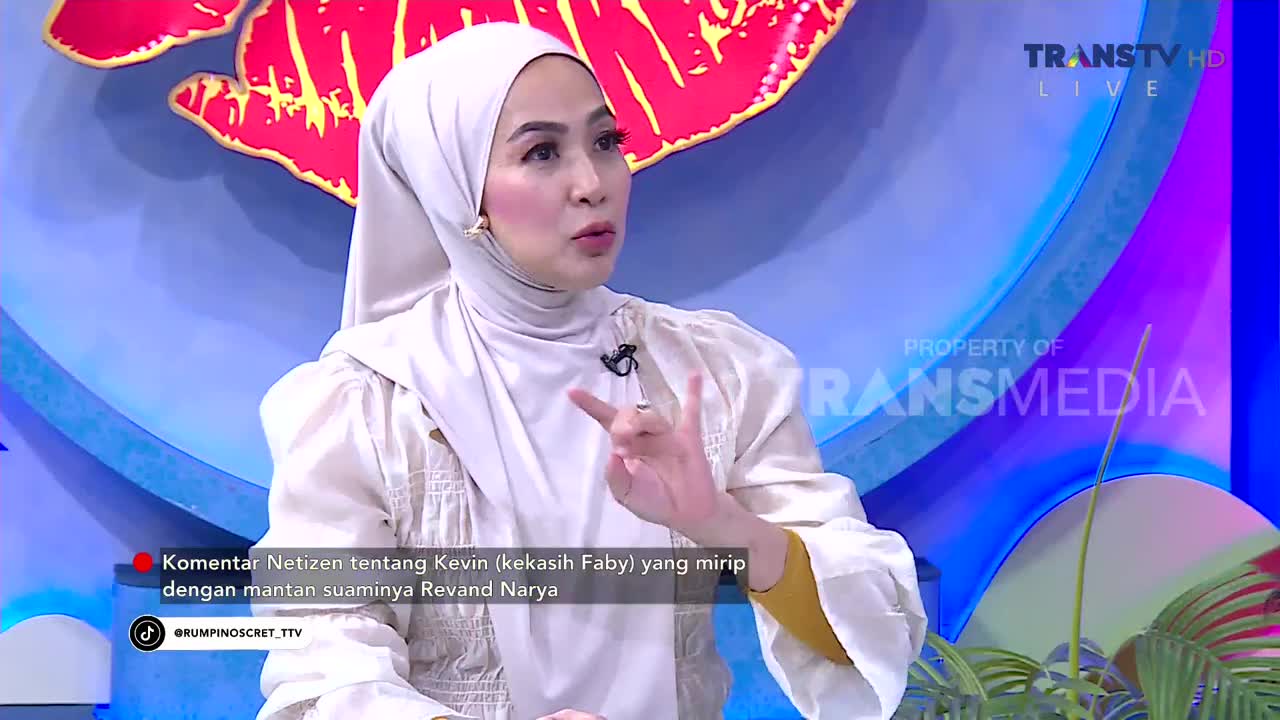 A woman in a white hijab speaks animatedly on a Trans TV set, gesturing with her hands. The background features a large, stylized red shape and the Transmedia logo.