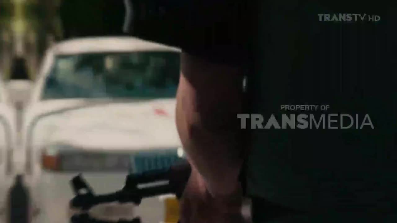 A man stands with what looks like an assault rifle, a yellow object in his hand, near a white car. The Trans TV logo is visible on the screen.