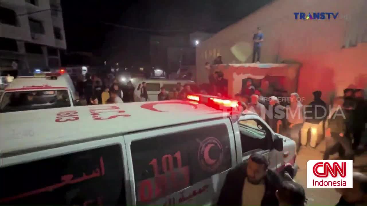 Red lights flash from the top of an ambulance as a crowd gathers. People stand on a low wall, watching the scene unfold.