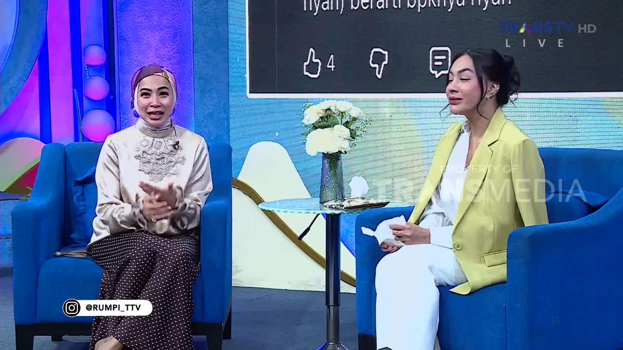 Two women sit on blue couches in a studio, one in a beige top and patterned skirt, the other in a yellow blazer and white pants. The Trans TV logo is visible behind them, and a bouquet of white flowers sits on a small table between them.