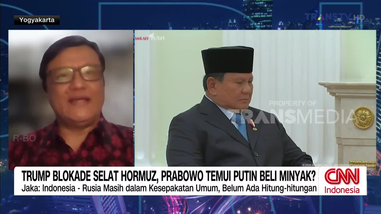 A man in a dark suit and traditional cap sits attentively, while another man in a patterned shirt speaks from a screen. A graphic at the bottom of the screen displays text in Indonesian.