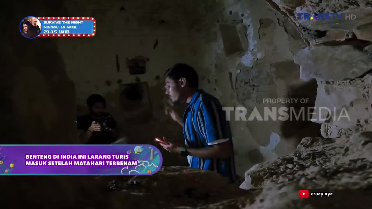 A man in a striped shirt shines a light into a dark, stone interior, holding his phone. Another person, mostly obscured, holds a camera in the background.