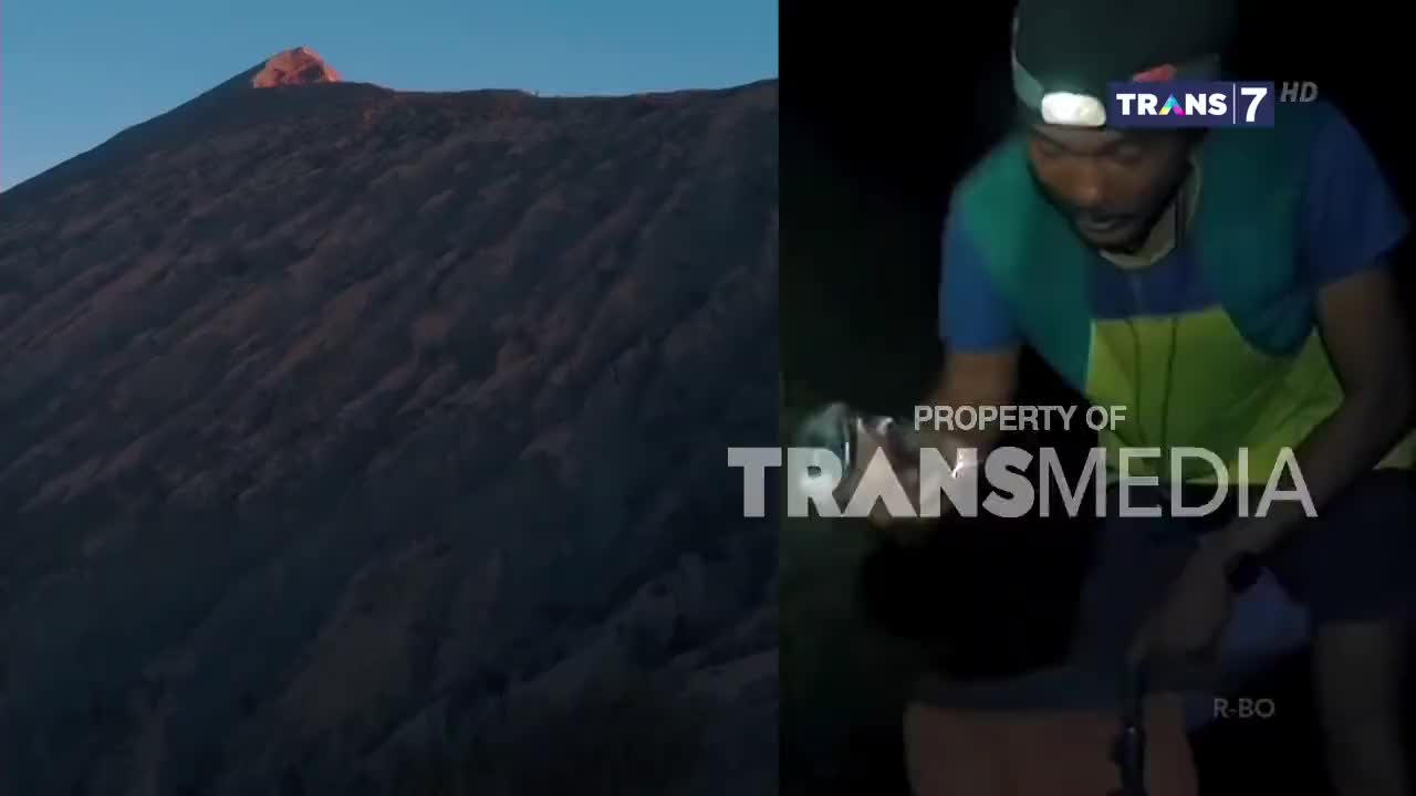 A mountain peak glows in the early morning light as a man, illuminated by a headlamp, examines something in his hands. The scene is being broadcast on Trans7 from Indonesia.
