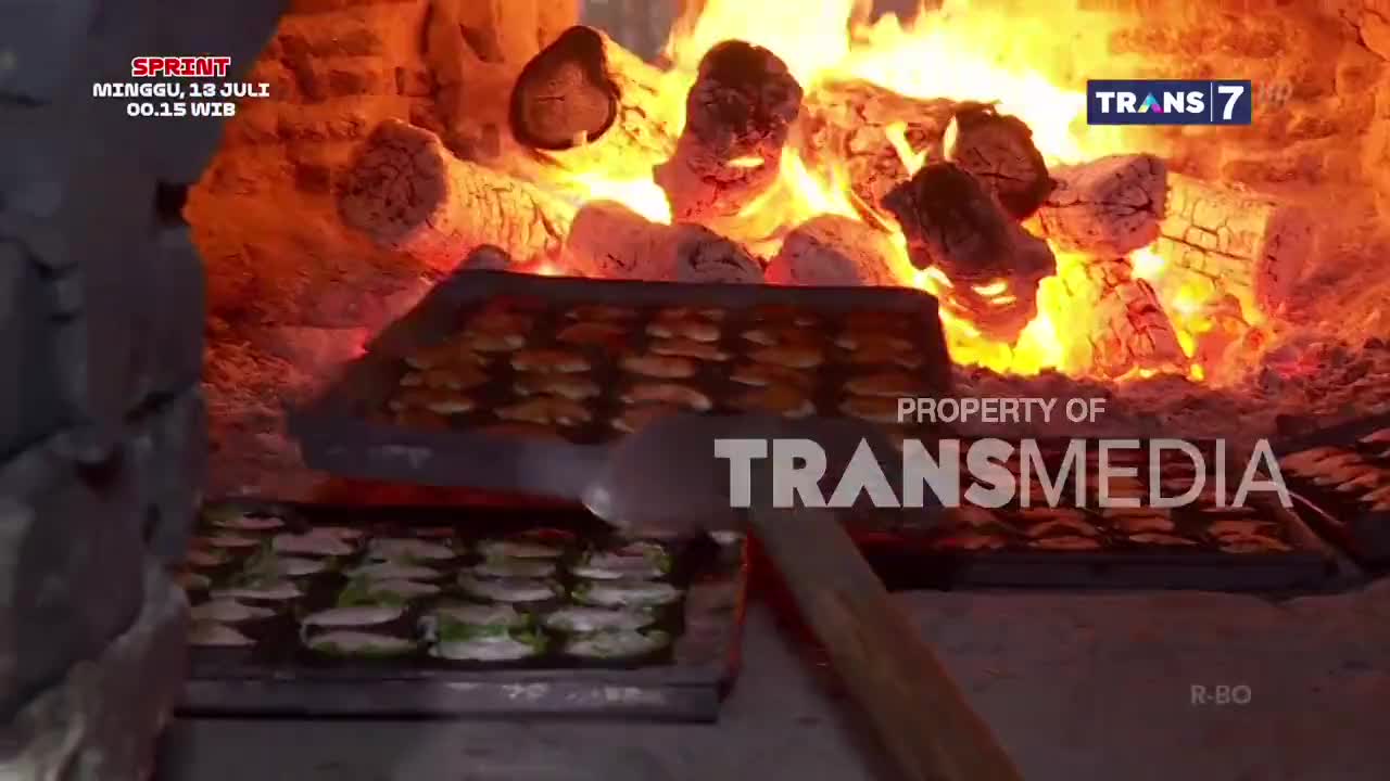 A tray of pastries is being pulled from a wood-fired oven, the flames casting a warm glow. The Trans7 logo is visible, suggesting this might be part of a cooking segment.
