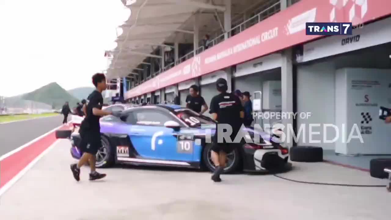 A race car, adorned with the number 10, is being attended to by a team of mechanics in front of a garage. The scene is broadcast on Trans7, with the Lombok International Circuit visible in the background.
