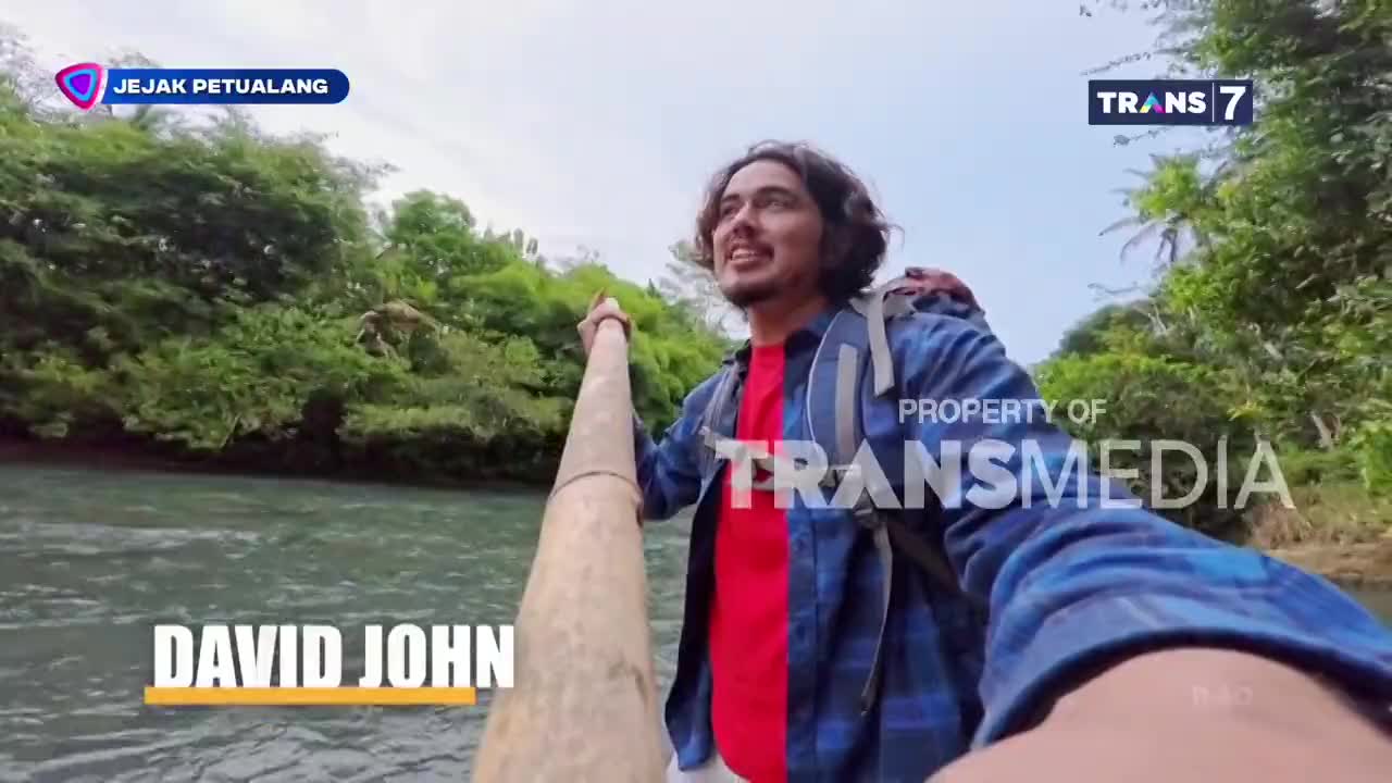 David John from "Jejak Petualang" on Trans7 is navigating a river, holding a long pole to guide the small craft. Lush green foliage lines the banks as he smiles, looking ahead.
