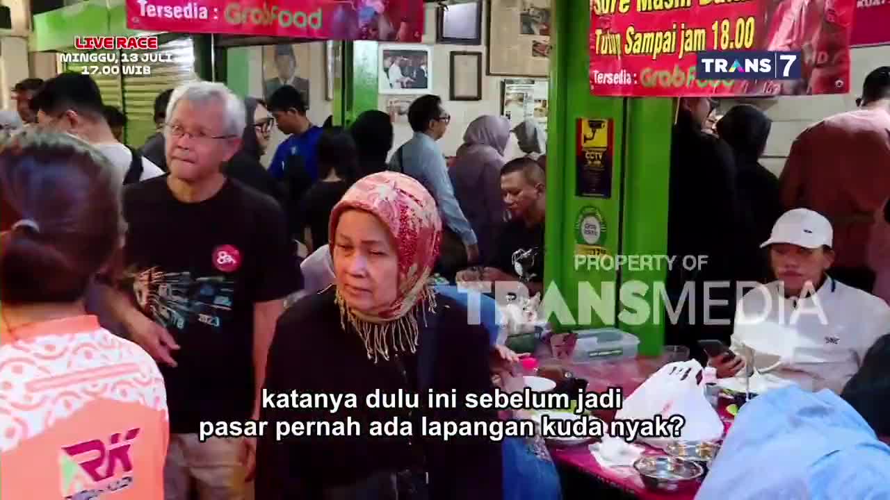People are gathered inside a crowded space, some seated at tables, others standing and talking. A Trans7 logo and the words "LIVE RACE" are visible, suggesting a broadcast is underway.
