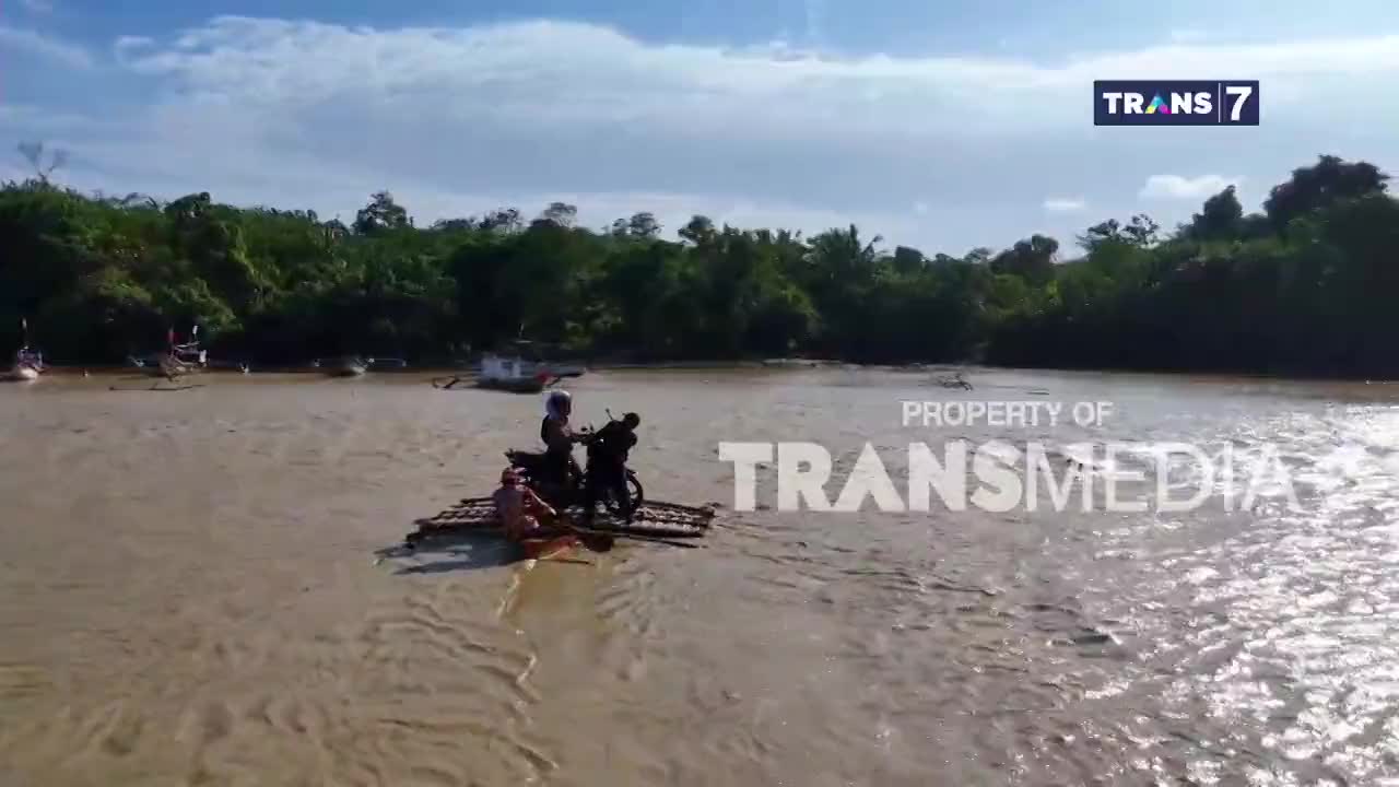 A motorcycle sits on a bamboo raft, navigating the murky water. Two people are paddling, while the rider sits upright, likely ready to disembark on the other side.

