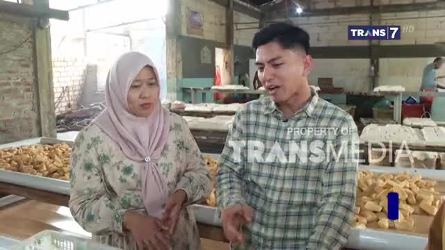 A woman in a headscarf and a man in a plaid shirt are gesturing, likely explaining something to the camera. Behind them, trays of food sit on long tables, and the Trans7 logo is visible in the corner.
