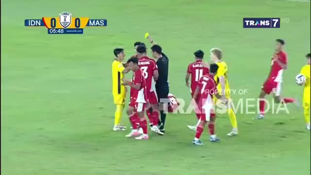 The referee is holding up a yellow card, and the Indonesian players in red are arguing. A player in yellow stands nearby, watching the scene unfold on the Trans7 broadcast.
