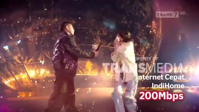 A man in a sparkly jacket holds out a microphone towards a woman, who covers her mouth in surprise. The Trans7 logo and an advertisement for IndiHome internet are visible in the background.
