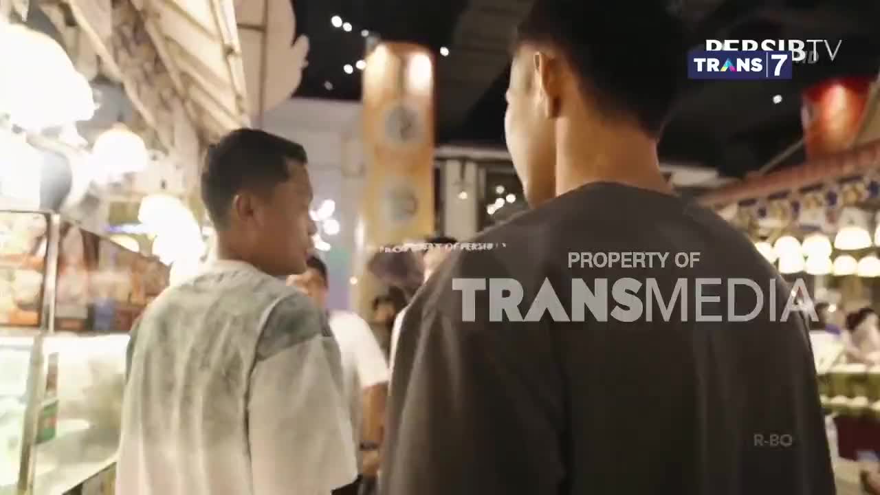 Two men walk through a brightly lit shopping area, their backs to the camera. The Trans7 logo is visible in the corner, suggesting this is a broadcast from Indonesia.
