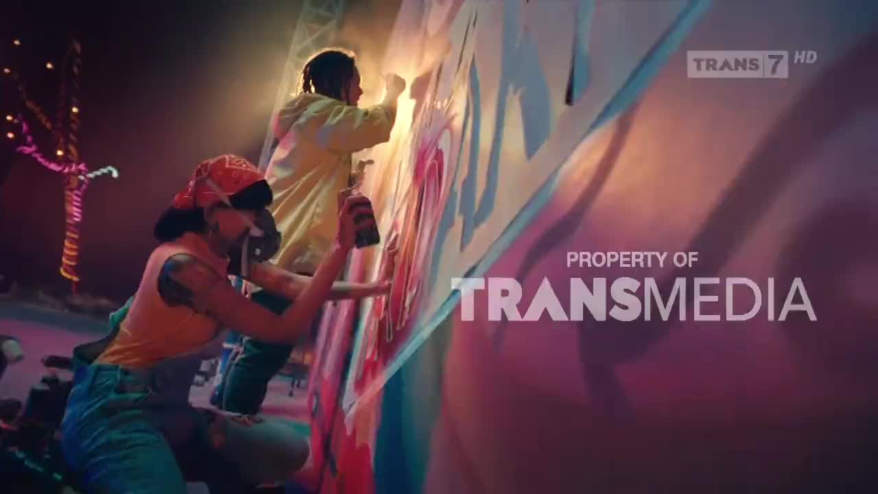 Two people are spraying a large wall with vibrant colors, likely for a Trans7 production. One person, in a yellow jacket, is working higher up, while the other, wearing a bandana, sprays from a lower angle.
