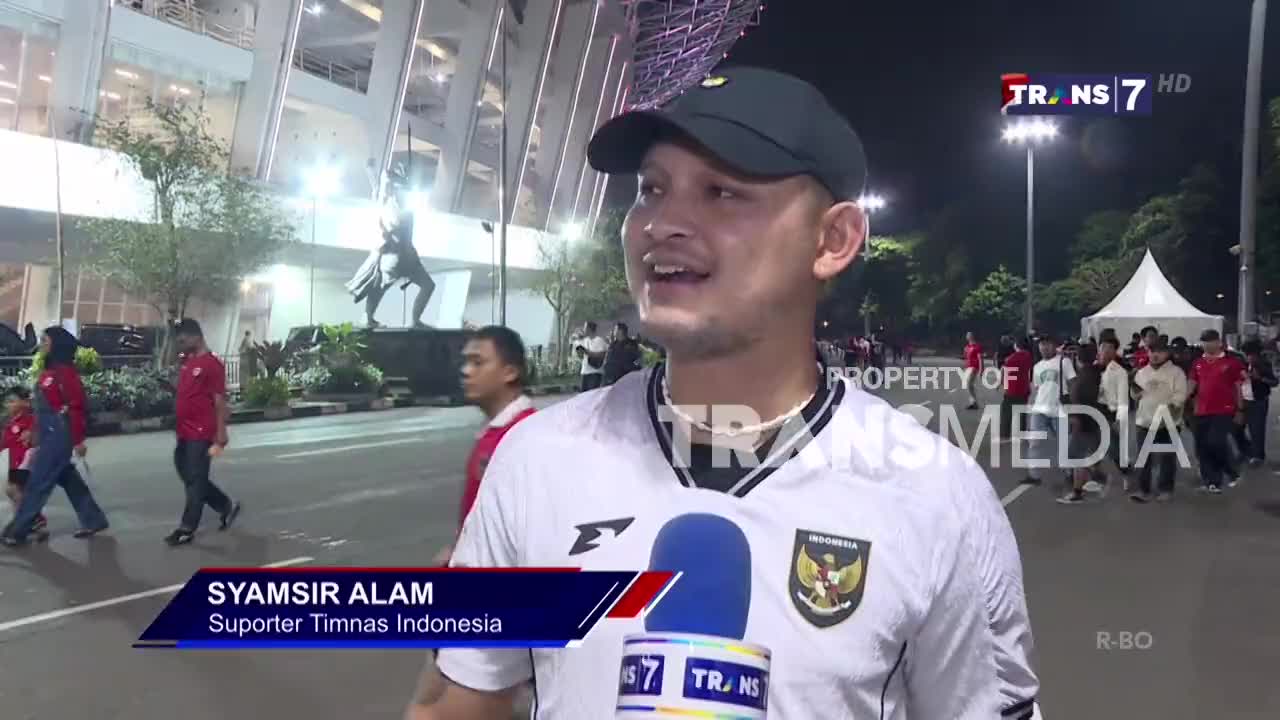 A man in a cap is being interviewed by a Trans7 reporter, with the Indonesian team's crest visible on his jersey. Behind him, a crowd of supporters in red shirts and white jerseys are walking towards the stadium.
