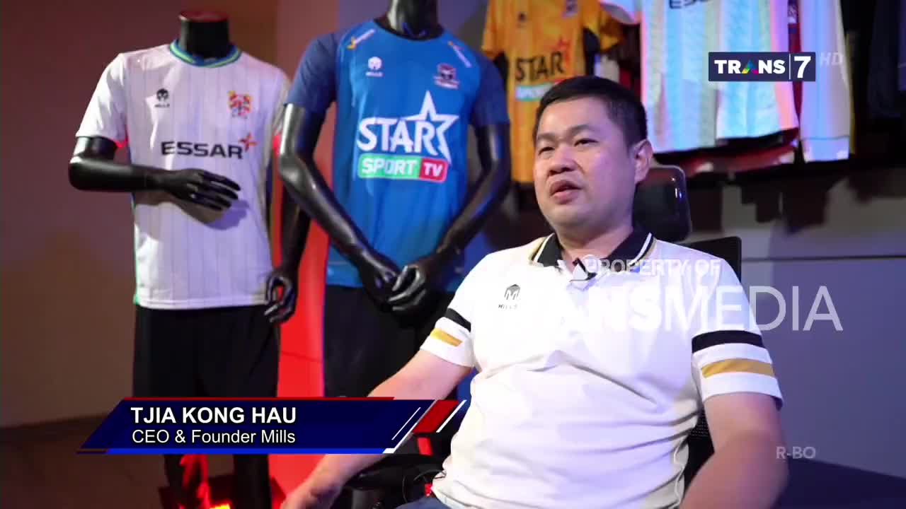 Tjia Kong Hau, the CEO of Mills, is seated and speaking on a Trans7 set. Behind him, mannequins display new soccer jerseys, with the "ESSAR" logo visible on one.
