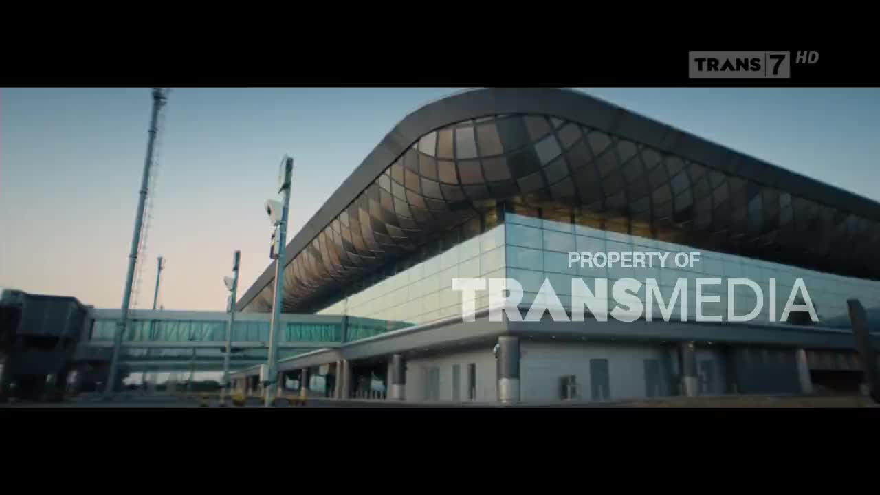 The camera is focused on a modern building with a glass facade, likely an airport terminal. The "Property of Transmedia" graphic is prominently displayed, and the Trans7 logo is visible in the corner.
