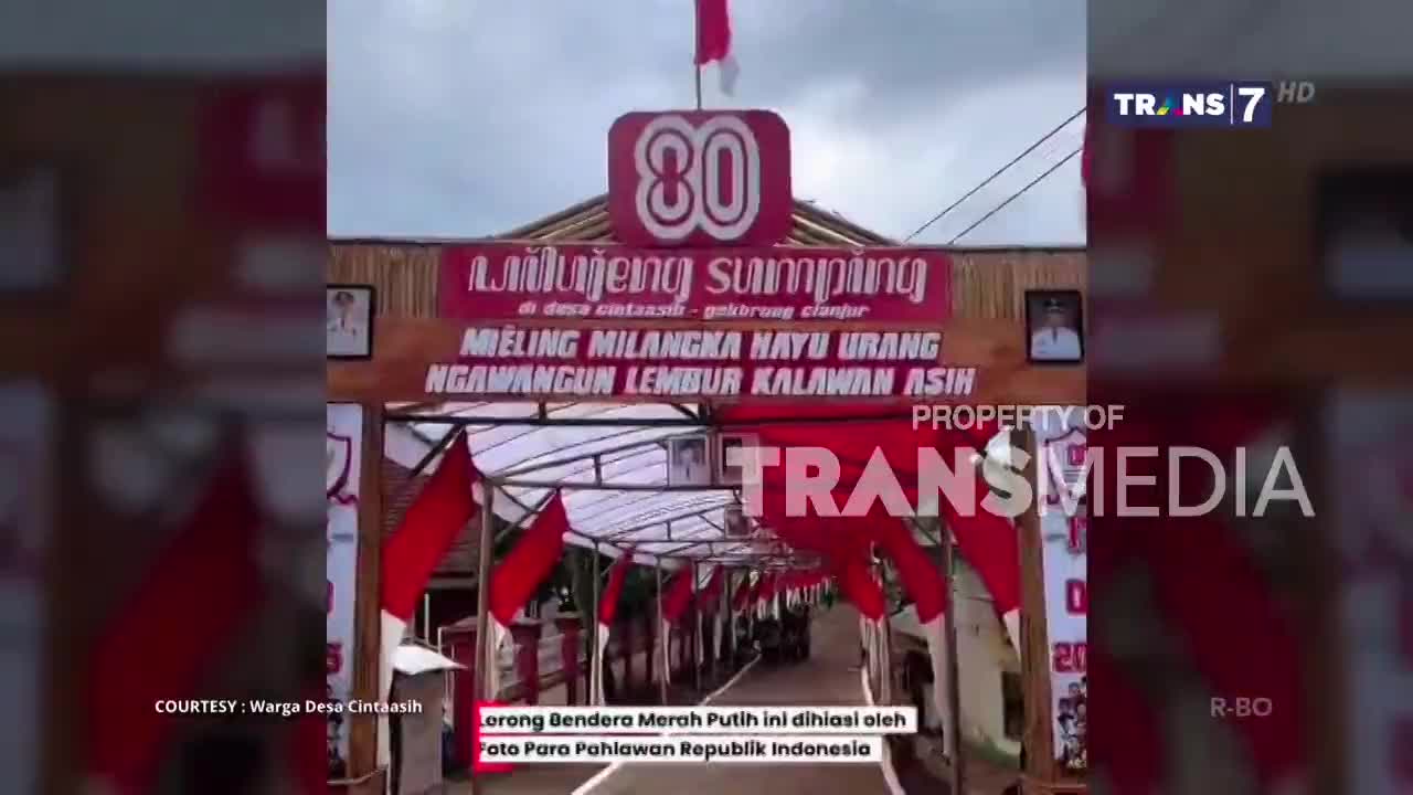 A decorated archway, celebrating an 80th anniversary, frames the entrance to a village. Red and white flags hang from the structure, and the Trans7 logo is visible in the corner.
