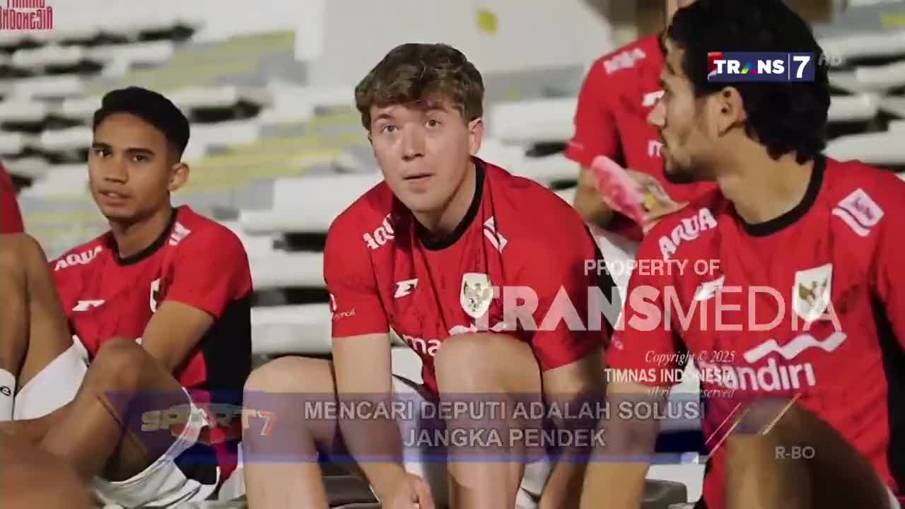 Players in red jerseys sit on a bench, some looking directly at the camera. A Trans7 logo is visible in the background, along with text in Indonesian.
