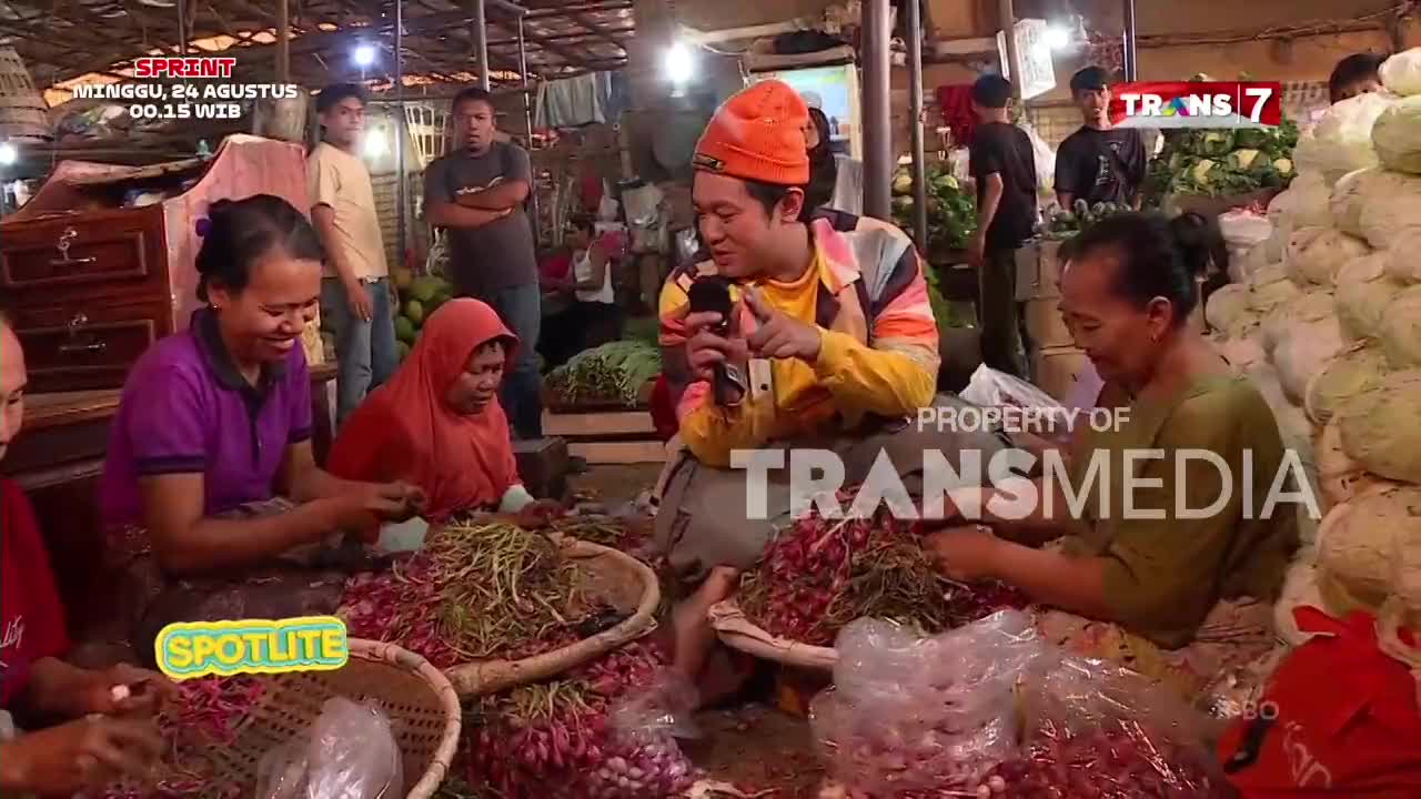 A man in a bright orange hat and jacket is holding a microphone, speaking to the camera while surrounded by women sorting through piles of red onions. The scene is part of a Trans7 broadcast, with "SPOTLITE" visible in the corner.
