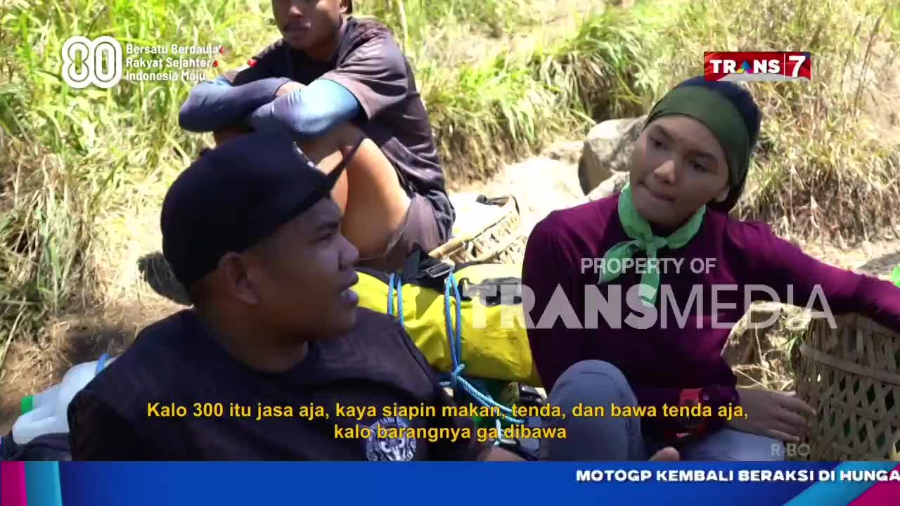 Three people are seated outdoors, engaged in conversation, with a Trans7 logo visible. The person in the foreground is speaking, while the others listen, seemingly discussing something related to camping or supplies.
