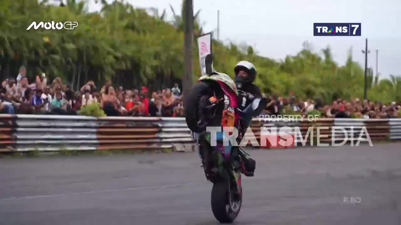 A motorcycle, its front wheel high in the air, is balanced by a rider holding a sign. The crowd, visible behind the trackside barriers, seems to be watching the spectacle, likely from a Trans7 broadcast in Indonesia.
