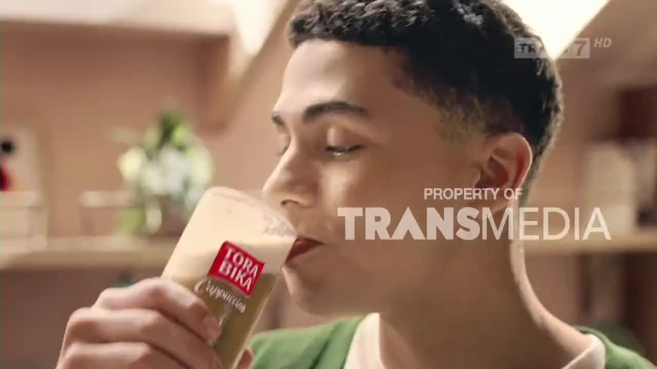 A young man is taking a drink from a glass of cappuccino, the Tora Bika logo visible on the side. The scene is likely part of a commercial, as indicated by the Trans7 and Transmedia branding.
