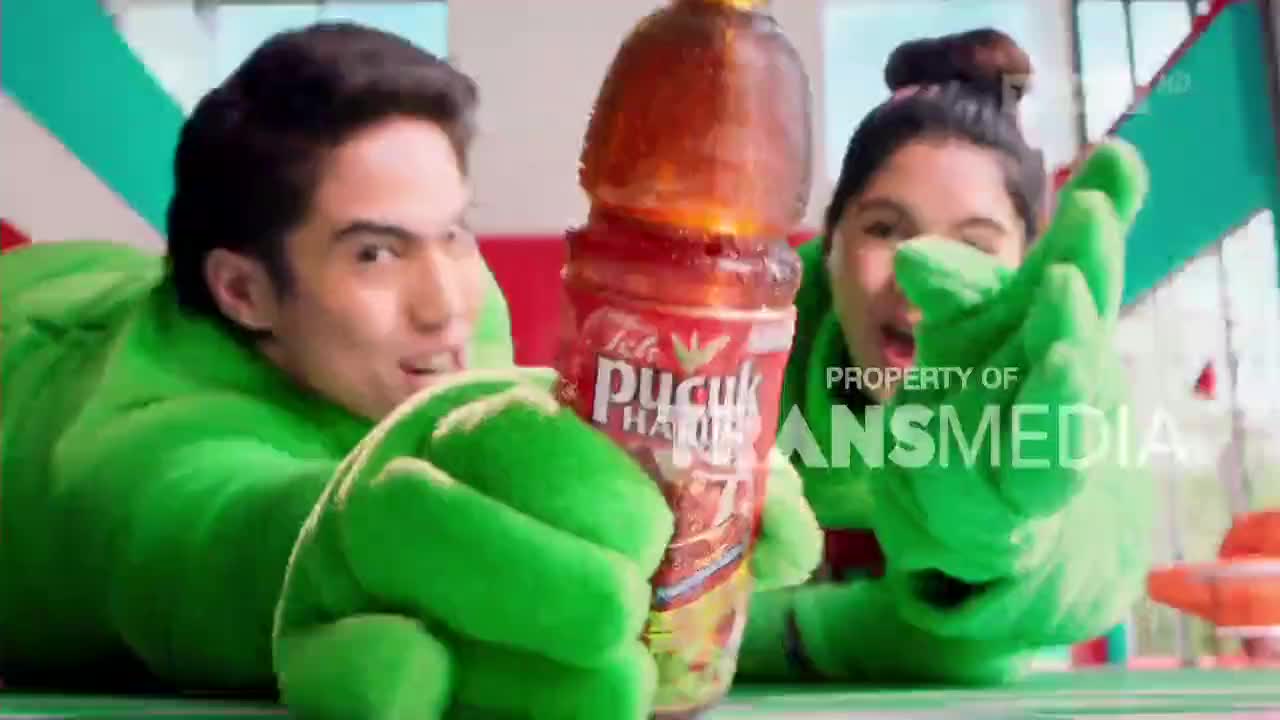 Two people in green, oversized suits are holding up a bottle of tea, grinning at the camera. The Indonesian brand, Teh Pucuk Harum, is clearly visible on the bottle.
