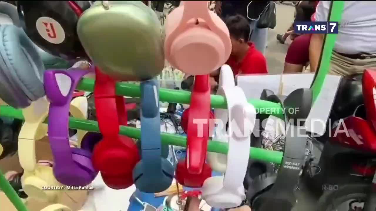 Headphones in various bright colors hang on a green rack, likely for sale. A person in a red shirt is visible in the background, possibly browsing the merchandise, as the Trans7 logo is displayed.
