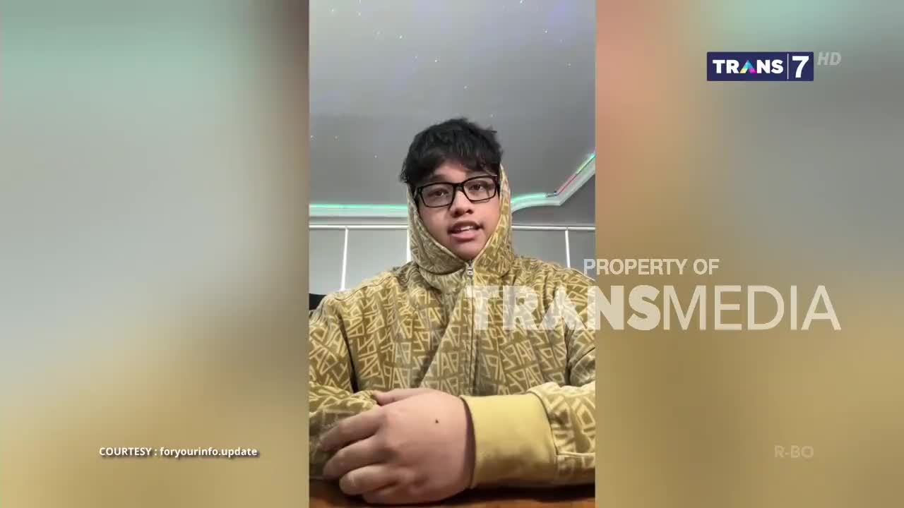 A young man in a patterned hoodie speaks directly to the camera, his face framed by the hood and glasses. The logo for Trans7, an Indonesian television network, is visible in the top right corner.
