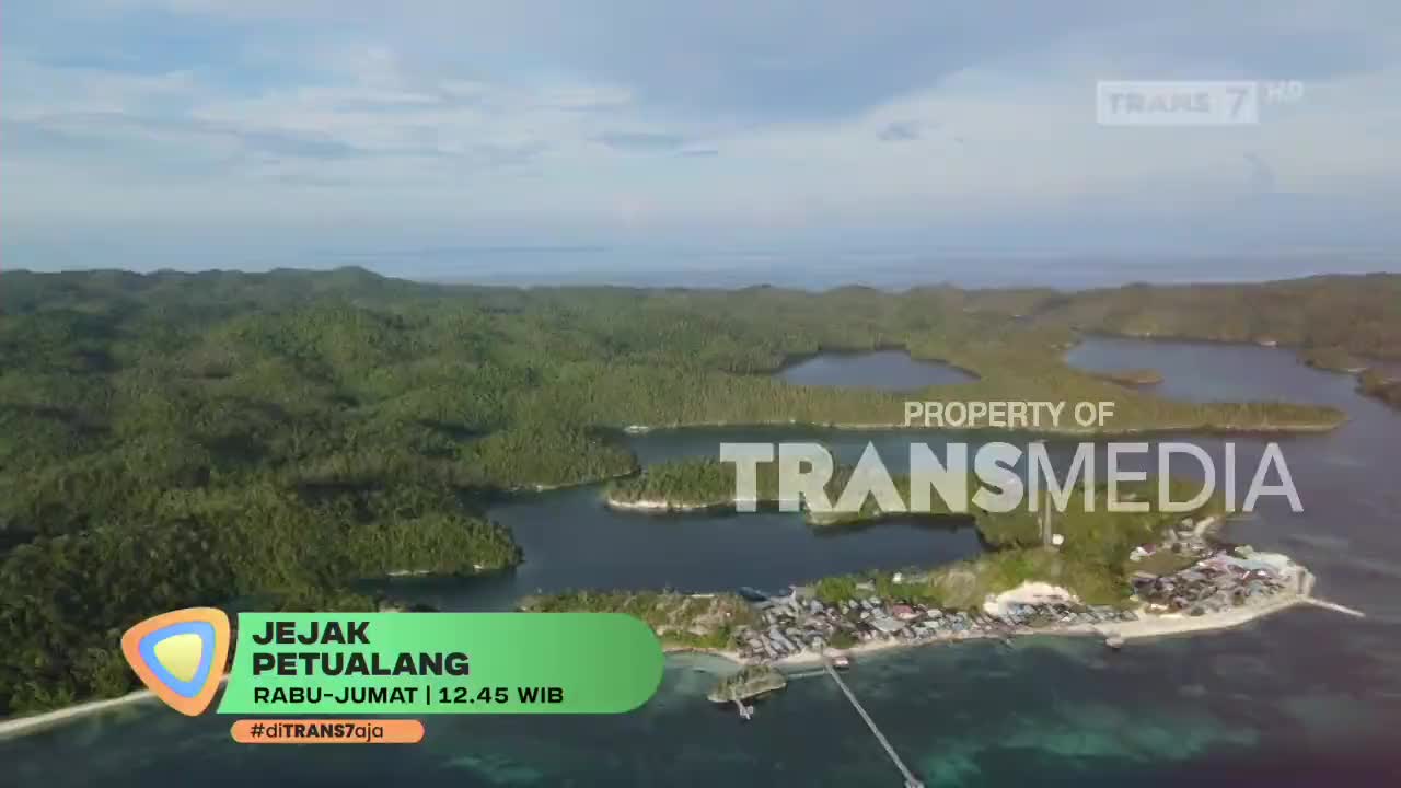From above, a lush, green landscape dotted with water is visible, with a coastal village at the bottom. The Trans7 logo and show title, "Jejak Petualang," are displayed on the screen.
