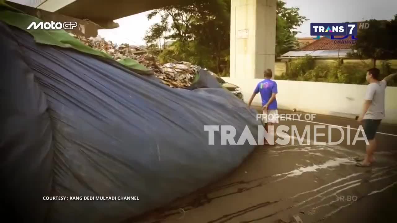 Underneath a bridge, a large, dark tarp covers a debris pile, and two men stand nearby. The Trans7 logo is visible in the corner, suggesting this is footage from Indonesia.

