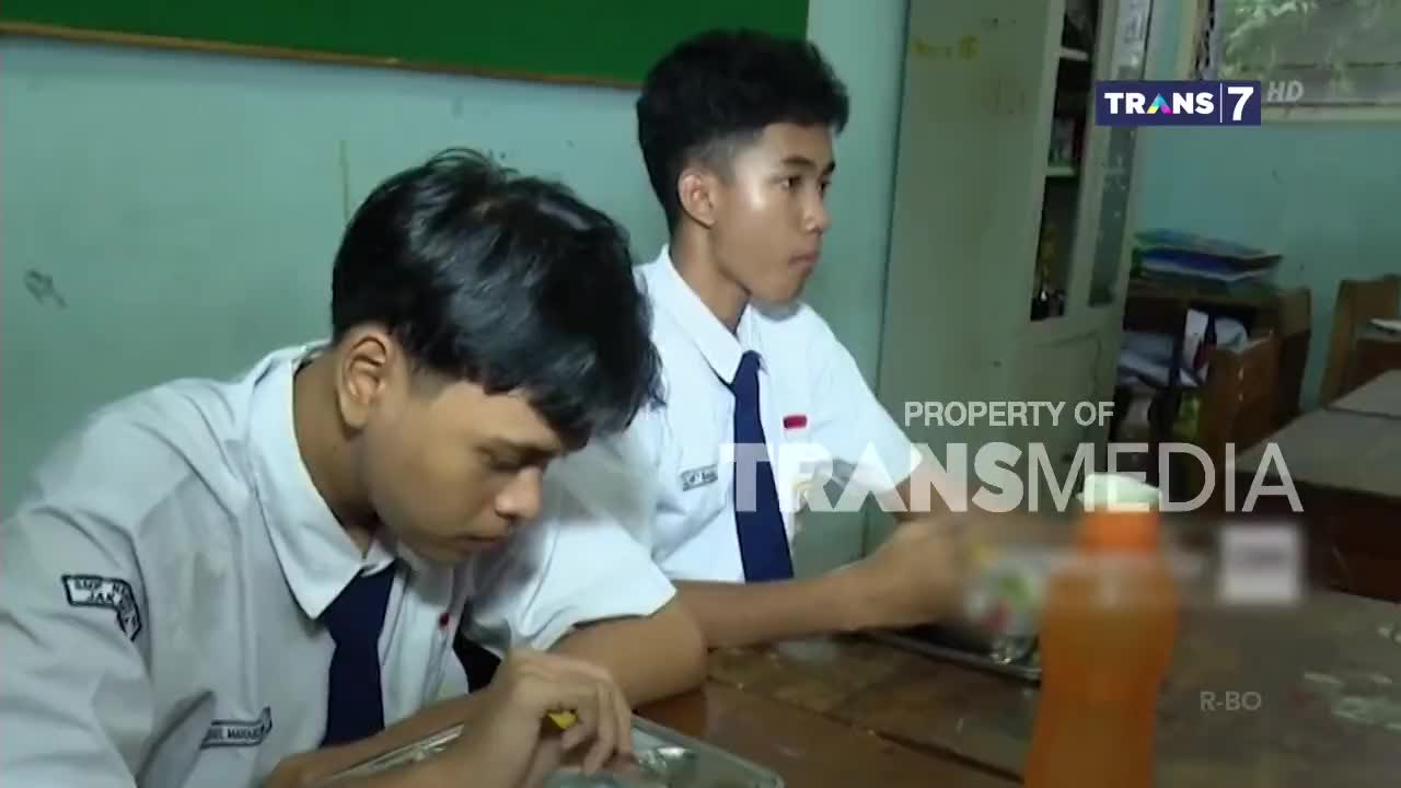Two students in white shirts and ties sit at a table, one looking down at a tray and the other holding a bottle. The scene is being broadcast on Trans7, as indicated by the logo.
