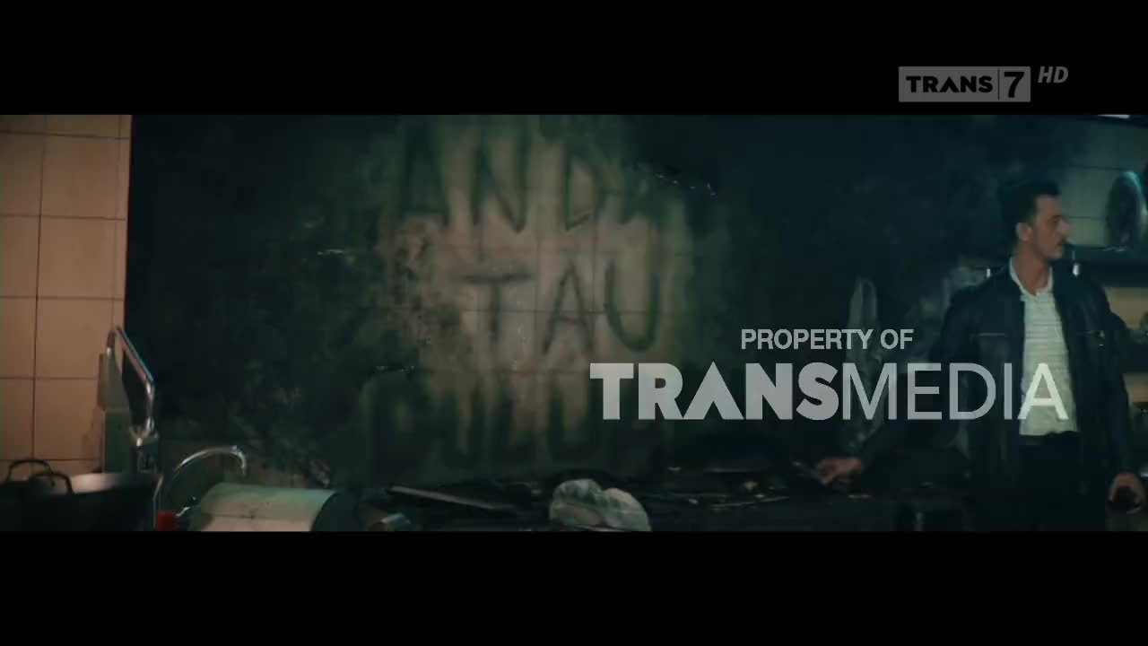 Smoke billows across a dark wall scrawled with Indonesian text. A man in a jacket stands near the edge of the frame, looking towards the graffiti, as the Trans7 logo appears.
