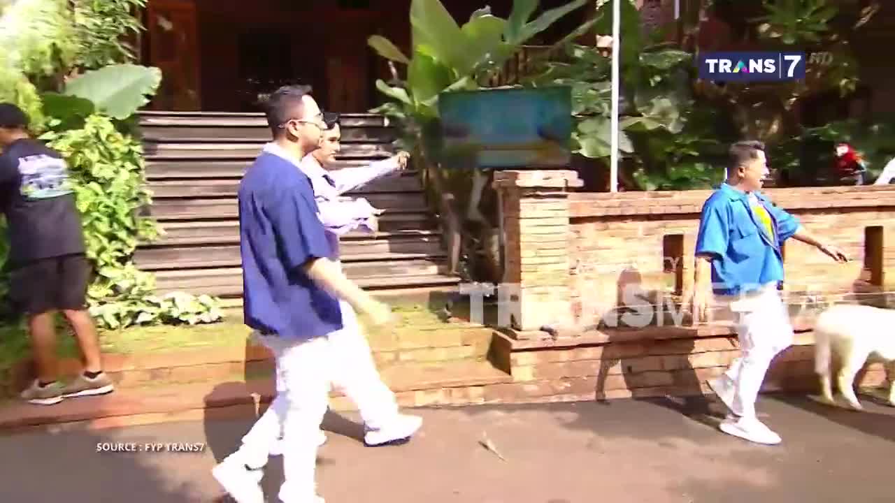 Three men in white pants and blue shirts walk down a brick-lined path, one with a white dog trailing behind. The scene is being filmed for Trans7, as indicated by the logo in the corner.
