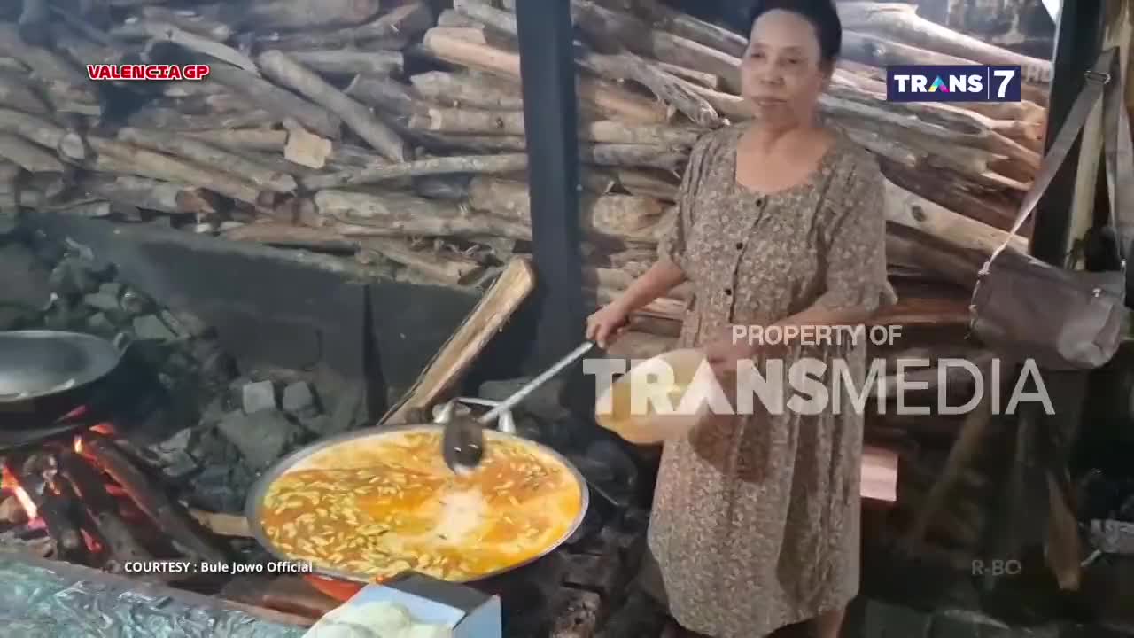 A woman stirs a large pot of simmering food over an open fire, a ladle in one hand and a bowl in the other. The Trans7 logo is visible, suggesting this is a cooking segment from Indonesia.
A woman stirs a large pot of simmering food over an open fire, a ladle in one hand and a bowl in the other. The Trans7 logo is visible, suggesting this is a cooking segment from Indonesia.