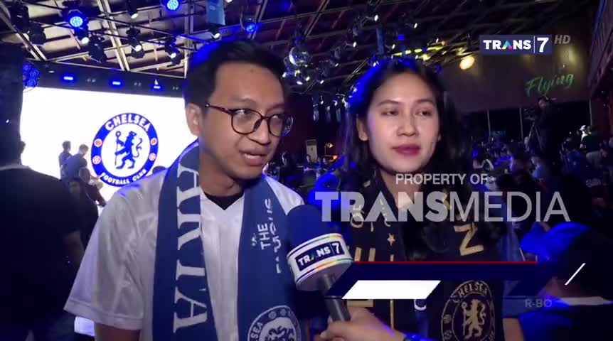 A man and woman, both wearing Chelsea scarves, are being interviewed by a Trans7 reporter. Behind them, a large Chelsea logo is displayed on a screen, with a crowd of people in the background.
A man and woman, both wearing Chelsea scarves, are being interviewed by a Trans7 reporter. Behind them, a large Chelsea logo is displayed on a screen, with a crowd of people in the background.