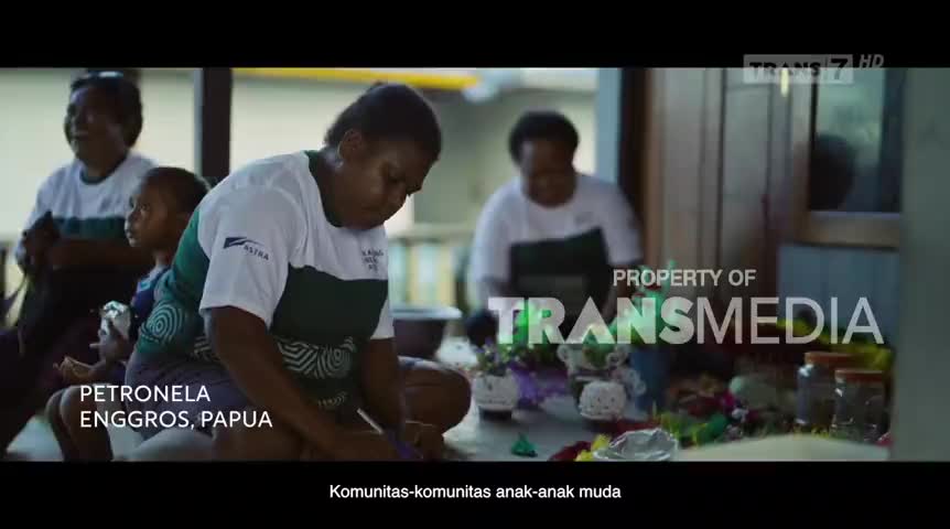 A woman in a green and white shirt sits on the floor, focused on something in her hands. Other women and children are nearby, and the scene is being filmed by Trans7 in Papua, Indonesia.
A woman in a green and white shirt sits on the floor, focused on something in her hands. Other women and children are nearby, and the scene is being filmed by Trans7 in Papua, Indonesia.