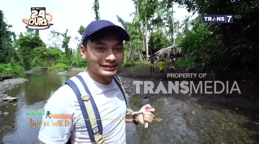 The man in the cap is wading through the shallow water, facing the camera for Trans7. Further up the stream, two figures are walking near a makeshift shelter, likely for the "Into the Wild" segment.
The man in the cap is wading through the shallow water, facing the camera for Trans7. Further up the stream, two figures are walking near a makeshift shelter, likely for the "Into the Wild" segment.