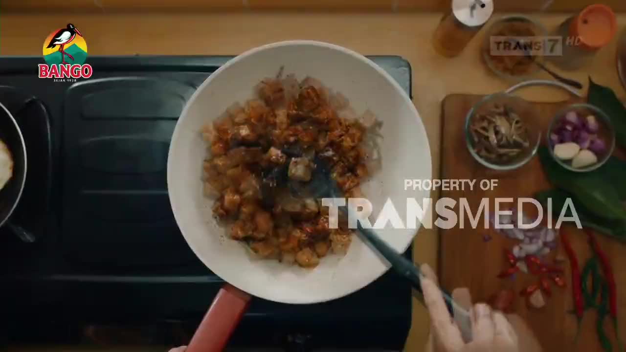 A spatula stirs cubed tempeh in a white pan on a black stove. Nearby, chopped shallots and chilies lie on a wooden cutting board.