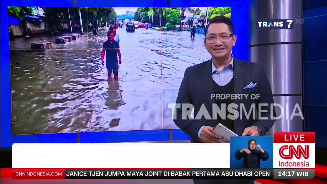 A man wades through knee-deep water on a flooded street in Indonesia, while a Trans7 presenter smiles on screen. Cars are visible in the background, submerged up to their wheels.