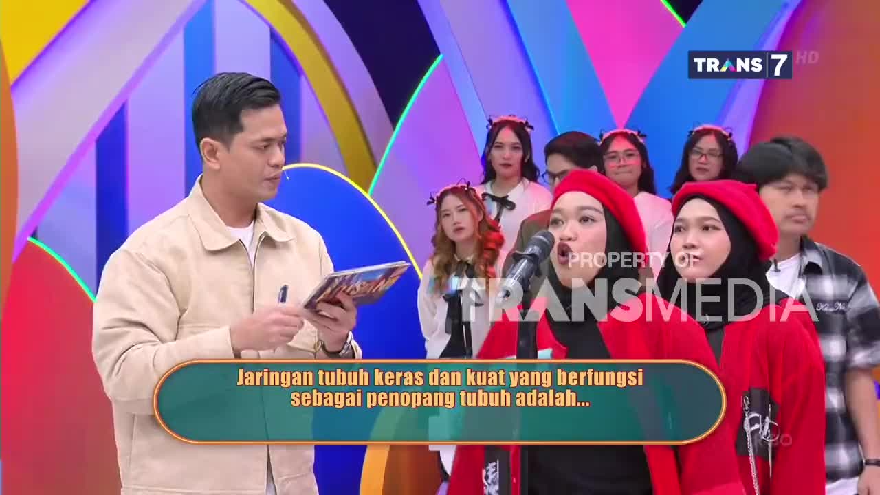 The host on Trans7 holds a tablet, looking towards the two young women in red. They sing into a microphone, their faces animated.