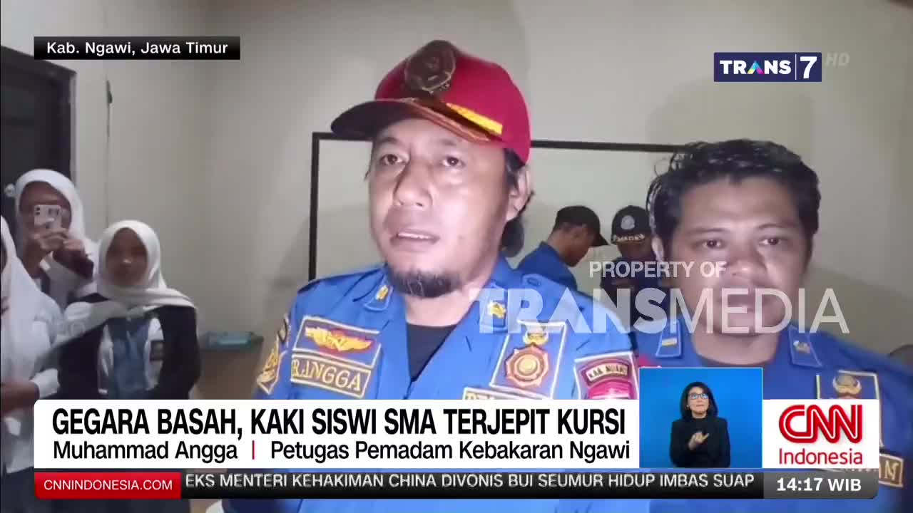 A man in a blue uniform with "RINGGA" on his sleeve speaks directly to the camera, while behind him, students in white uniforms and headscarves stand by. Another man in a similar uniform stands to his right, looking forward.