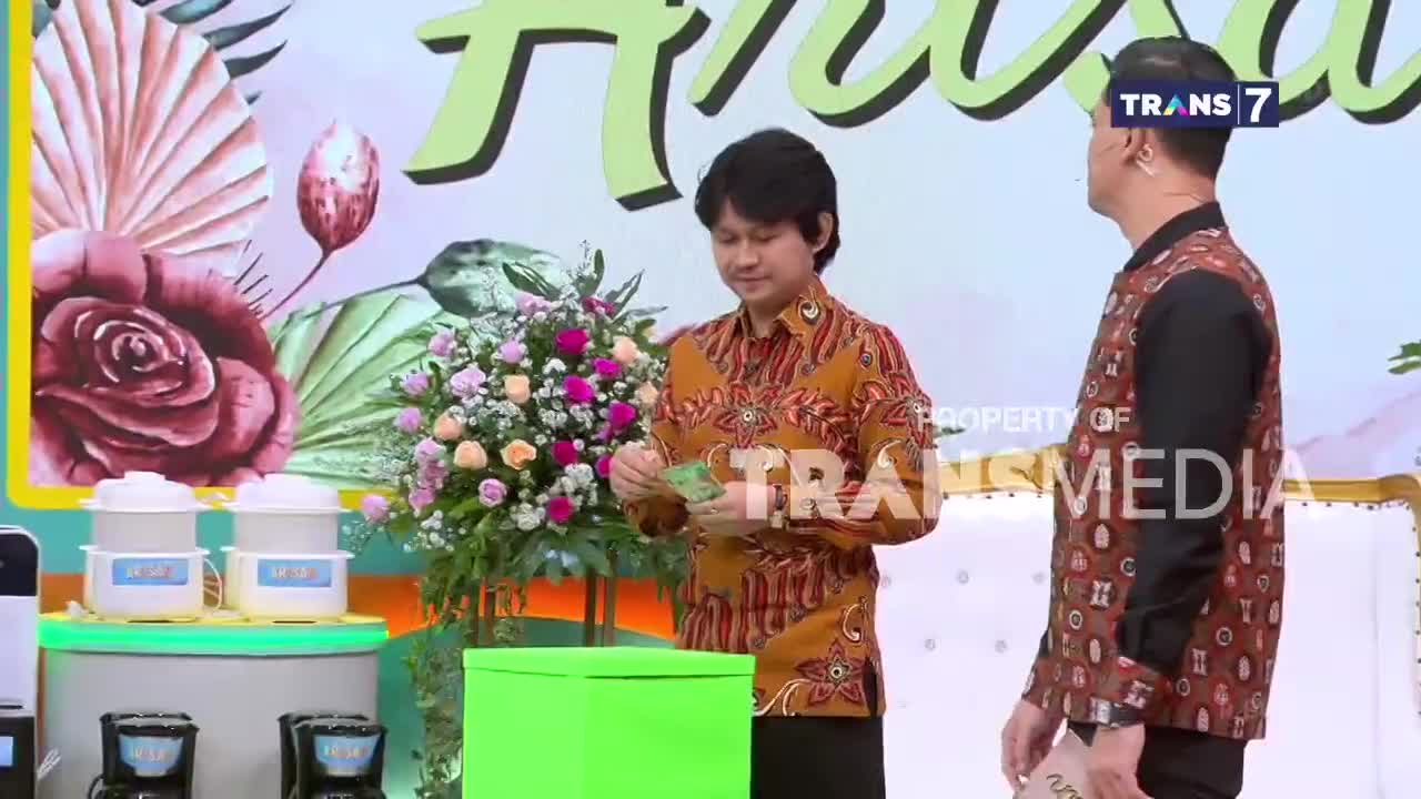 A man in a batik shirt holds out some Indonesian Rupiah bills, preparing to place them into a bright green box. Another man stands nearby, facing away, his patterned vest a striking contrast to the floral backdrop. This is happening live on Trans7.