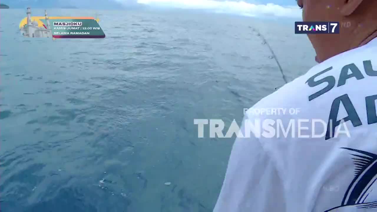 The ocean rolls under a cloudy sky as a fisherman, wearing a shirt with "SAUL AD" printed on it, casts his line. A graphic for "MASJIDKU" and the Trans7 logo are visible on screen.