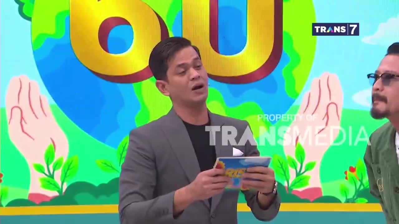 The host on Trans7 gestures while holding a colorful tablet. Behind him, a large graphic of the earth and cupped hands fills the screen.
